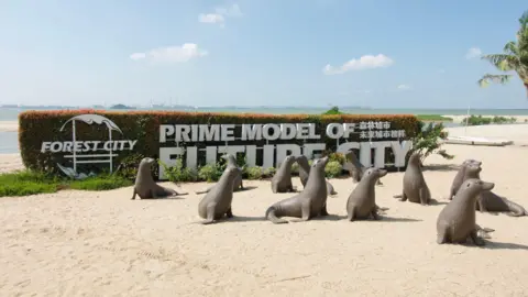 BBC/ Gavin Butler Twelve seal statues on a each in front of a hedge sporting a white "Forest City" logo and the words "Prime model of future city" in all capitals. Ocean in the background.