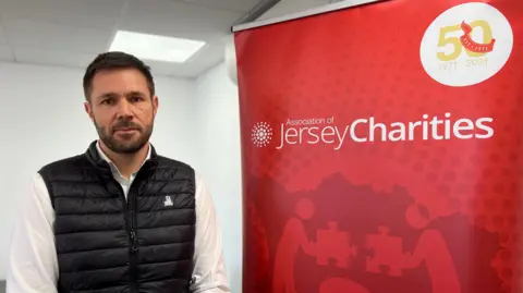 A man with short dark hair and a dark beard. He is wearing a black puffa-style gilet over a white shirt. He is staring directly at the camera, to the left of the image. On the right of the image is a red, floor-to-ceiling banner, which says 'Association of Jersey Charities'. In the top right corner of the banner, it says '50' in large gold numbers, above smaller numbers which say 1971-2021.