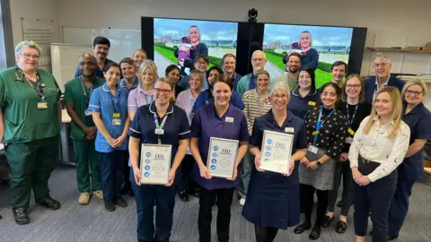 UHNM A team of people - some wearing medical scrubs - stood in a group, smiling while posing for the camera. Three of the group, stood at the front, are holding framed certificates.