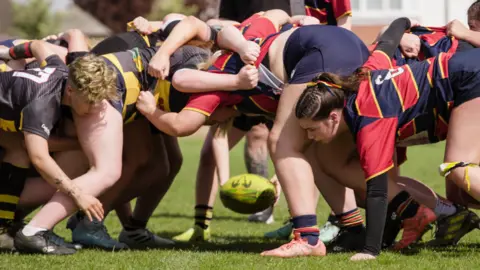 O Museu e a Galeria de Arte de Northhampton são um Scrami em uma partida de rugby feminina. Um grupo de vermelho e azul está voltado para o outro em amarelo e preto. Uma bola amarela está prestes a ser mantida no scame.