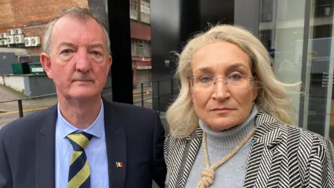 A man and a woman stand outside a building. He has grey hair and wears a blue shirt and jacket and a striped tie. She has blonde hair and glasses and wears a grey jumper and black and white striped jacket with a thick necklace around her neck.