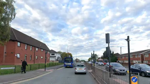 A street view of a busy main road with cars either side and terraced houses and flats alongside it. A person in black walks past a road sign for Birchfield Lane carriageway.