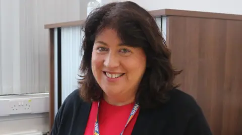 University Hospitals Dorset NHS Foundation Trust Siobhan Harrington smiles at the camera. She has shoulder-length, brown, wavy hair and wears a black cardigan over a red top with an NHS lanyard.