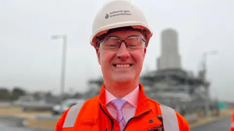 Emma Baugh/BBC A man smiling at the camera in a picture taken with a blurred view of part of a gas plant behind him. He is wearing a white hard hat with the words "National Gas transmission" printed on it. He is also wearing a hi-vis orange jacket, a white shirt and a pink tie. He is wearing protective goggles.
