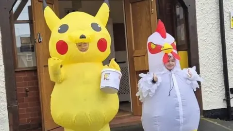 Family Two young boys standing outside. One is wearing an inflatable Pikachu costume, and the other boy, who is smaller, is wearing an inflatable chicken costume.