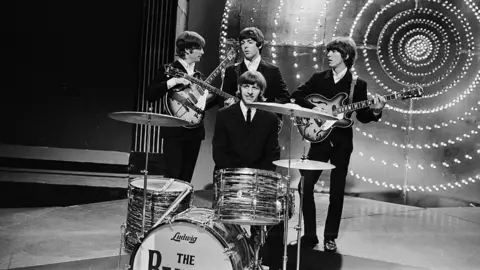 The Beatles pictured during rehearsal for appearance on BBC-TV's 'Top Of The Pops'. Standing behind drummer Ringo Starr are left to right: John Lennon, Paul McCartney and George Harrison. They are wearing suits and holding guitars. It is the early days and they all look young. 