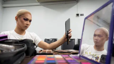 Getty Images Gawdland, Thai winner of Ru Paul's Drag Race UK versus the World, looking at the mirror while doing her make up with a wide array of makeup colours in the foreground