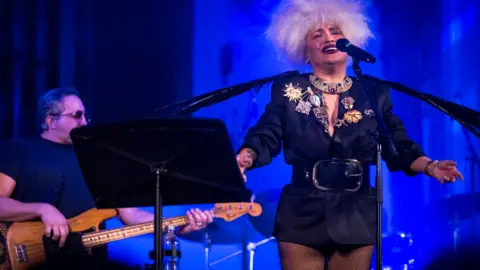 Still Moving Media A woman on a darkened stage in front of a mic, performing with her eyes closed and her arms outstretched. She is wearing a mini black jumpsuit with a huge buckled belt and lots of large brooches. She has a mop of unruly white hair. There is a guitarist behind her and a large black music stand in the foreground.