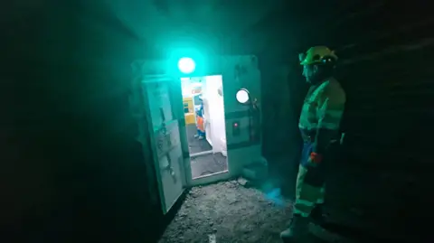 A worker in hi-vis clothing stands in front of the emergency pod. It has a strong light on the top. The door is opened and we can see it is lit inside.