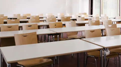 An empty classroom with beige single desks and a window in the corner of the room with blinds.