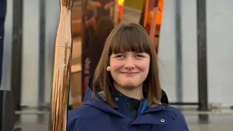 Millie Knight/King’s College London A woman smiles in front of the Paralympic Flame, she is holding a gold torch which is also alight.
