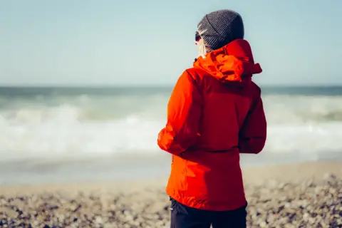 A woman wearing a red waterproof coat and a wooly hat with sunglasses stands with her hands in her pockets on a beach, looking out to the sea