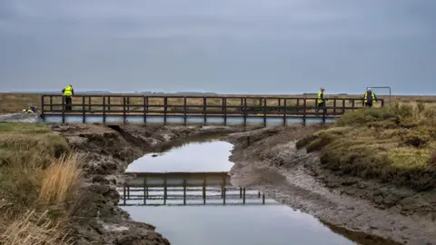 A general view of Stiffkey bridge - a footbridge crossing a marshland creek. There are three workers on the bridge.