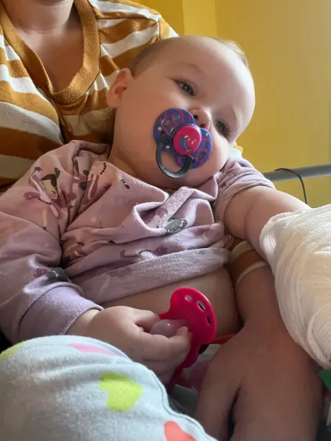 Family handout. A baby around ten months old is lying in her mother's arms looking unhappy. She is holding a dummy and has another in her mouth. The baby has a bandage on her arm and looks to be in a hospital bed. 
