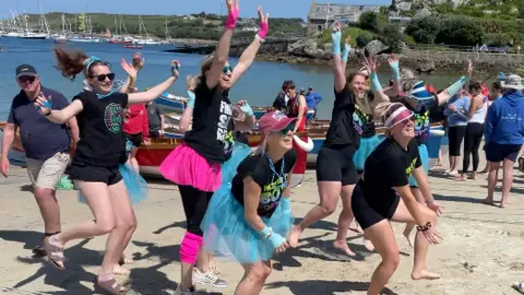 A group of people in fancy dress including tutus jump in the air on a beach.