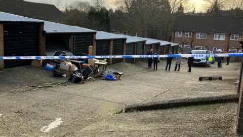 Louise Parry/BBC Police tape prohibits entry to some garages. Household items are stacked up outside one of the garages, and a police van and several uniformed and plain-clothes staff are in the distance. There are houses beyond the van