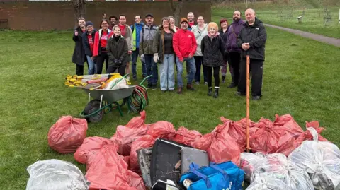 Friends of People's Park A large group of people, outside, wearing coats and warm clothing, in front of a large selection of bagged up rubbish. There is a wheelbarrow with equipment on it, a path to the right and large areas of green grass. 