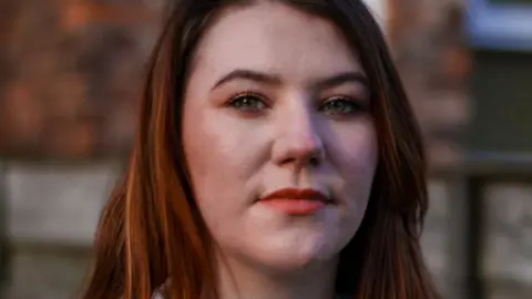 A still image of Jodie Morrow. She is staring at the camera, while she is stood against a wall outside. She has long, light coloured hair and is wearing a blue, knitted cardigan over a white t-shirt.