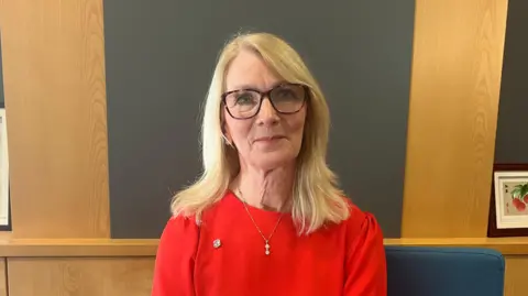 Professor Shirley Congdon, who has long fair hair and is wearing glasses. She is wearing a red jumper and has a necklace on. Behind her is wooden panels. 