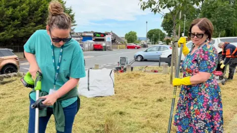 A woman in a turquoise T-shirt and black sunglasses trims grass, while another woman in a flowery dress rakes the grass.