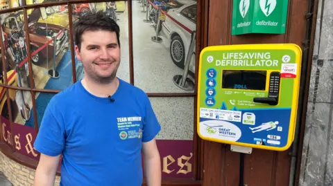 A man in a blue t shirt with short dark hair is stood next to a defibrillator. He is smiling, and directly behind him is a shopfront with pictures of cars in the window.