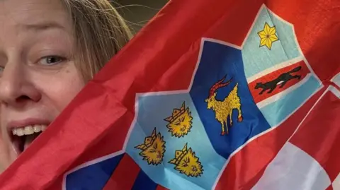 Emma Gale Emma Gale with an excited expression and her mouth wide, peeping from behind a Croatian flag. Part of her face is obscured.