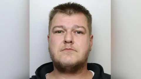 A man in a black hoodie with a earring in his left ear looks at the camera as part of a police mug shot