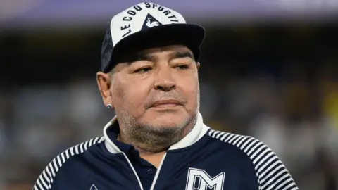 Getty Images Diego Maradona wears a navy and white cap and matching jacket at a football match in Buenos Aires. He has his right ear pierced and is looking away from the camera.