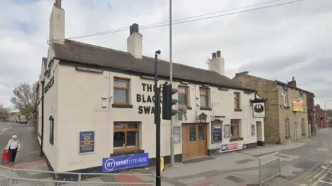 Google Front of the Black Swan pub with cream-coloured rendering and light brown wooden window frames and doors. It lies a corner of two roads next door to a smaller stone building to the right of the picture.