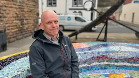 Federica Bedendo/BBC Kevin Kerrigan sat on a bench with mosaic decorations outside of Christ Church in Maryport. He is bald and has blue eyes. He is slightly smiling at the camera.