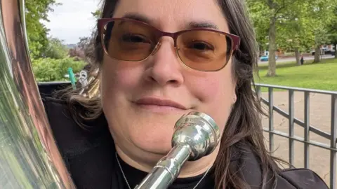 Rose Norman A woman with long black hair and sunglasses is holding a euphonium close to her mouth. She is stood outside near a park.