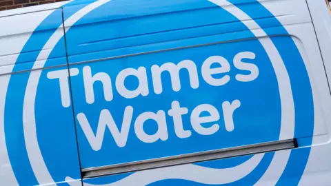 Blue and white Thames water logo on a van. It is close up. The logo is a blue circle with Thames Water written in white writing. There is a white wave below the writing.