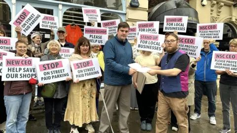 People are holding signs with pleas to stop parking charges. In the foreground, two men are handing over a paper with signatures.