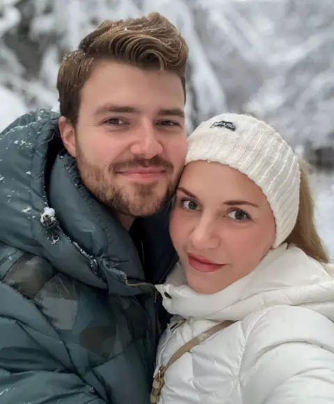 Handout Two people standing close together in the foreground, wearing padded winter jackets, with one in a dark green coat and the other in a white coat and knitted headband. Snow is visible on the clothing. Behind them are snow-covered trees and branches.