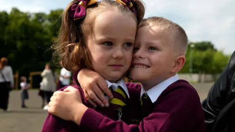 Young boy hugs his twin sister, they are wearing a red and yellow school uniform