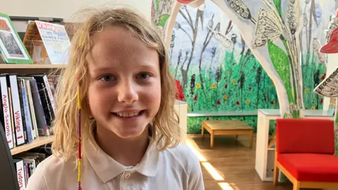 A young girl with blonde wavy hair is standing in her school library wearing a white polo shirt.