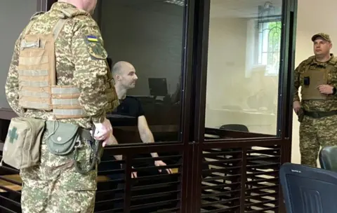 Slidstvo.Info  A man stands in a witness box in court with a soldier nearby