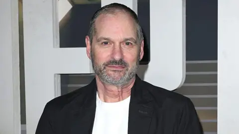 Getty Images A man wearing a black blazer and white t-shirt stares at the camera. He has short cropped greying hair and a well-trimmed grey beard.