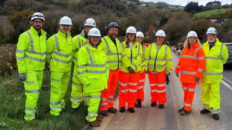 Anna Gelderd MP (fifth right) with National Highways and Octavius representatives on the recent site visit to the A38 Notter Bridge scheme. All ten of the people pictured are wearing his-vis jackets and trousers. They have white hard hats on. The group is made up of both women and men.