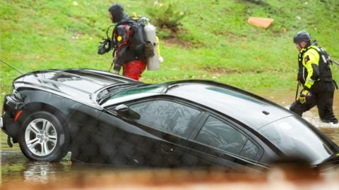 A police dive team walk past a car that is partly submerged in water