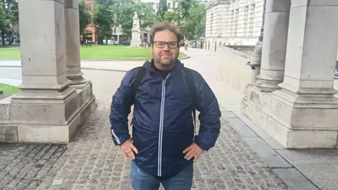 A man with glasses, brown hair and a thin brown beard stands with his hands on his sides. He is wearing a navy rain coat type jacket and jeans.
