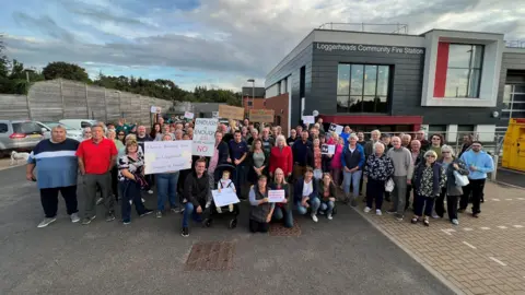A group of about 70 people holding banners are stood outside Loggerheads fire station. 