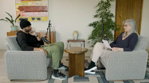 Made By Dyslexia A man on the left and a woman on the right are both sitting in chairs facing each other. They are both wearing dark tops. The woman has grey hair and is wearing glasses.