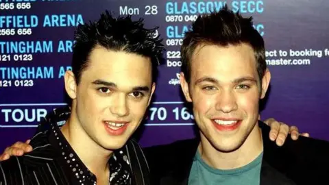 PA Media Two young men stand shoulder to shoulder smiling for a press launch, with a poster listing theatre dates behind them. The man on the left has black, spiky and gelled short hair and brown eyes and is wearing a black striped jacket and black shirt with white and grey spots. The man on the left has light coloured eyes, brown short spiky hair, a dark jacket and green tshirt. Each man has placed a hand on the other's shoulder 