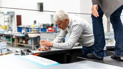 Ineos Britannia Geoff Willis with short white hair, wearing a grey sweater, leans on part of a grey hull in a test room. A pair of legs in blue trousers is visible to his left.