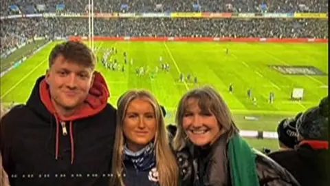 Family handout Liz Steele with son Paddy and another woman, standing in the crowd at a rugby game. Their backs are to the pitch as they smile for the camera.All three are smiling while wearing jackets and hoodies for the weather.