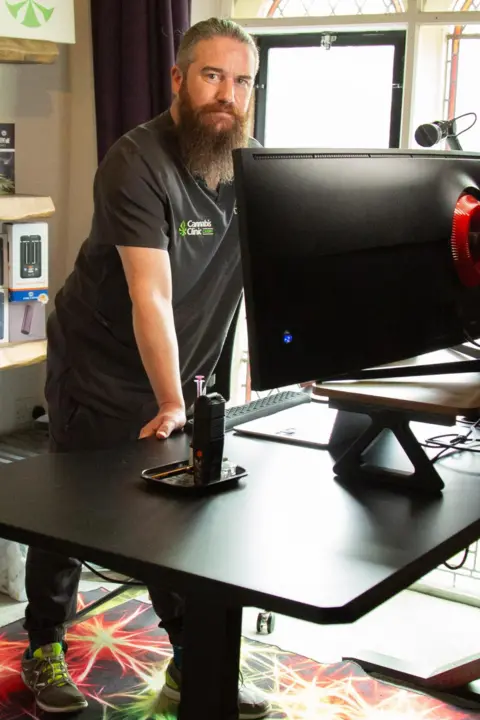 A man in scrubs with a long dark beard and tied back dark hair stand above a black desk in an office. Cannabis posters and medical books line the shelves behind him.