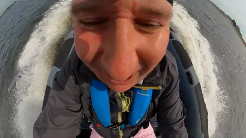 Wide angle close up of a man's smiling face, sitting in a speeding boat with wake and whitewater around the boat.