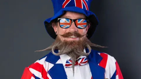 A man in a Union Jack hat, sunglasses, bow tie and suit, with a brown and grey beard shaped into five different points, two sticking to the side, one pointing down, two curling up