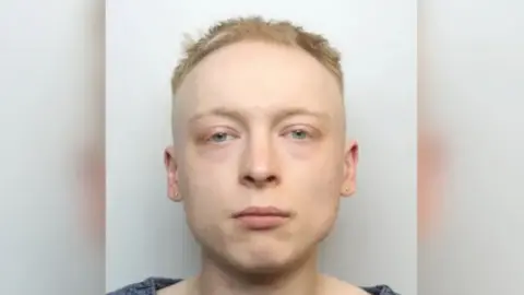 A mugshot of Miller, who has a vacant expression and is looking at the camera. He has short blonde hair and is wearing a blue top.